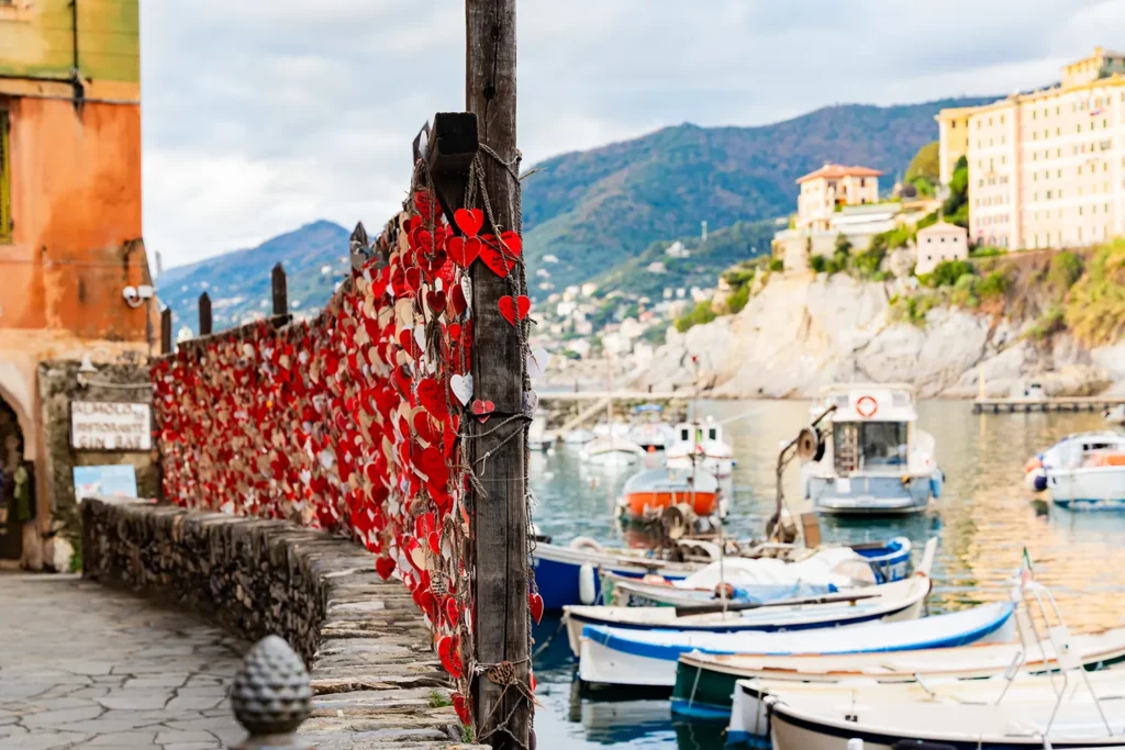 **Alt text:** Staccionata in legno lungo il porticciolo decorata con molti cuori rossi intrecciati, affacciata su barche da pesca ormeggiate; sullo sfondo case colorate e colline verdi che scendono verso il mare, in una tranquilla atmosfera costiera.